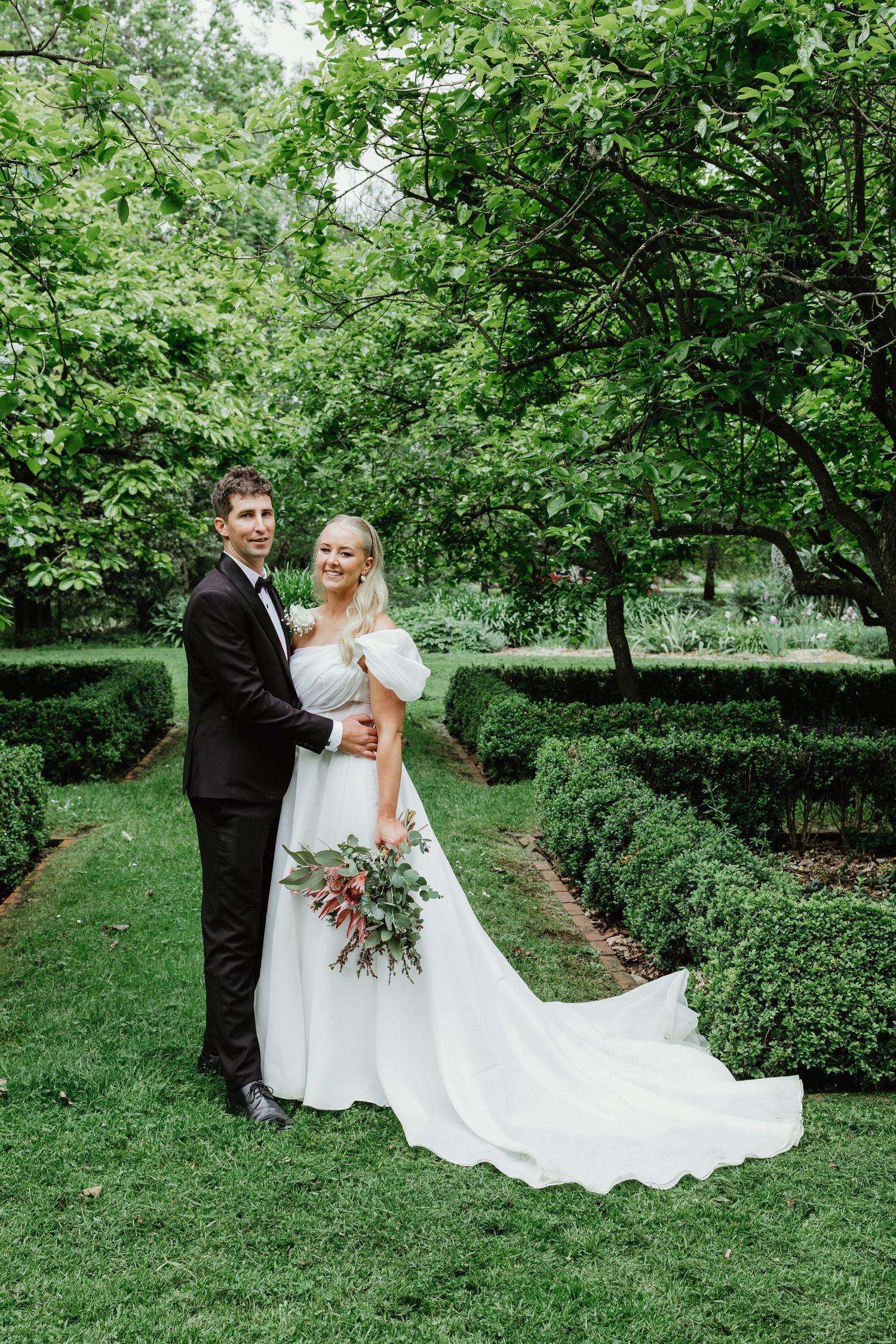 Bride and groom posing in lush green garden, bride in white gown with train holding bouquet, groom in black tuxedo.