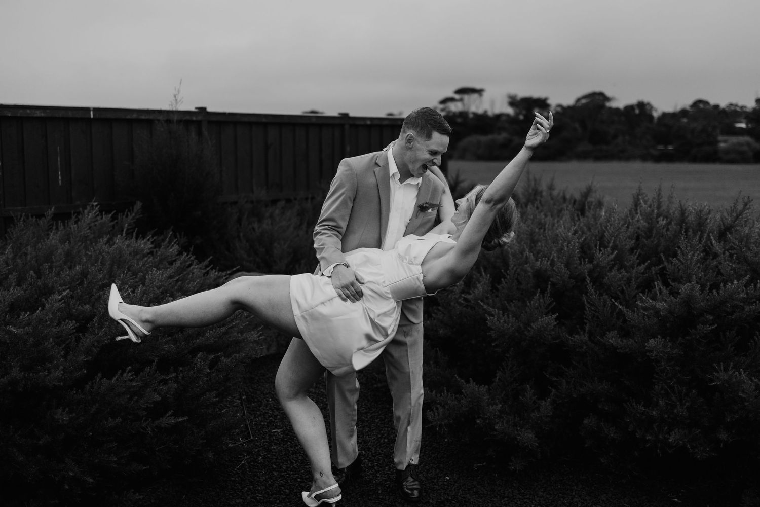 Black and white photo of groom dipping bride dramatically among garden shrubs at wedding.