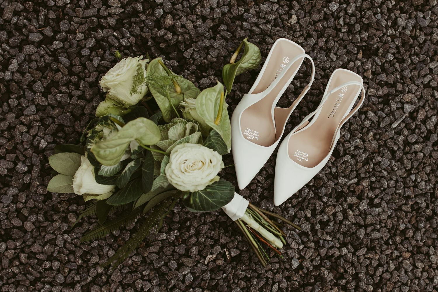 White bridal heels and green and white rose bouquet arranged on dark gravel for a wedding day flat lay.