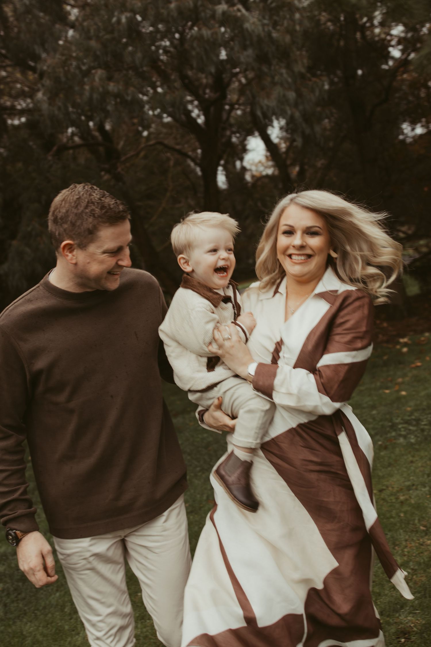 Happy family of three laughing and walking outdoors in coordinating brown and cream outfits.