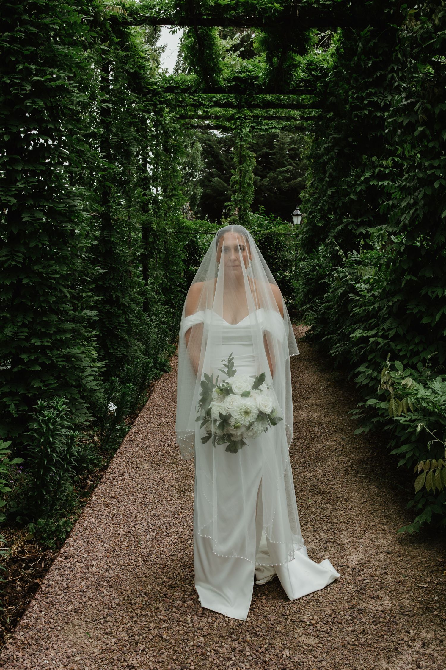 Bride in white gown and veil walks through a lush green garden archway holding a white bouquet.