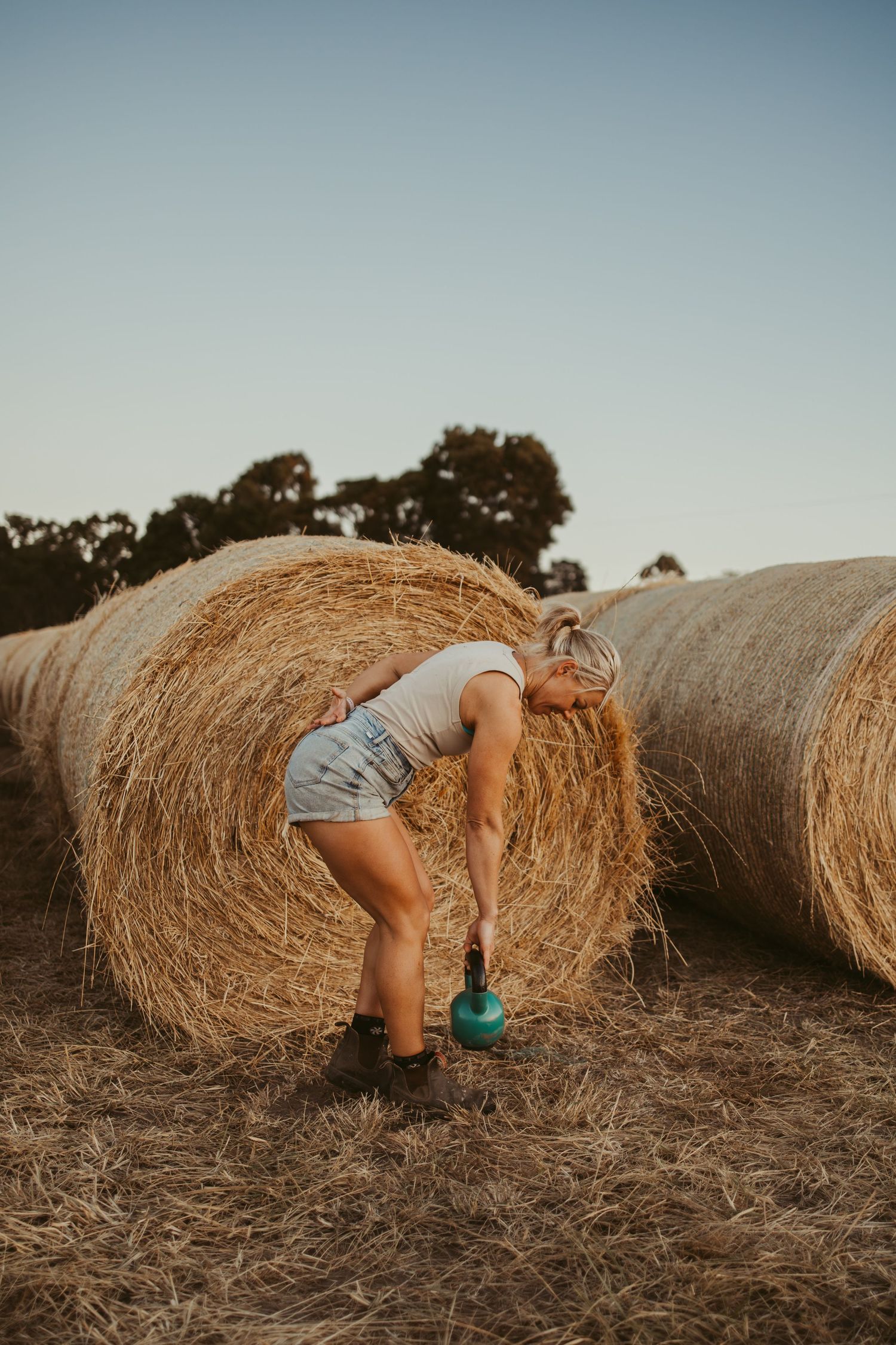 Woman doing kettlebell exercises in a field next to large round hay bales at sunset.