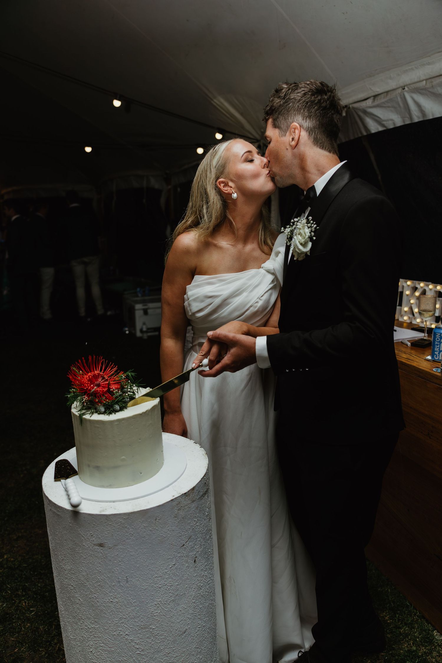 Bride and groom share kiss while cutting simple white wedding cake decorated with red protea.