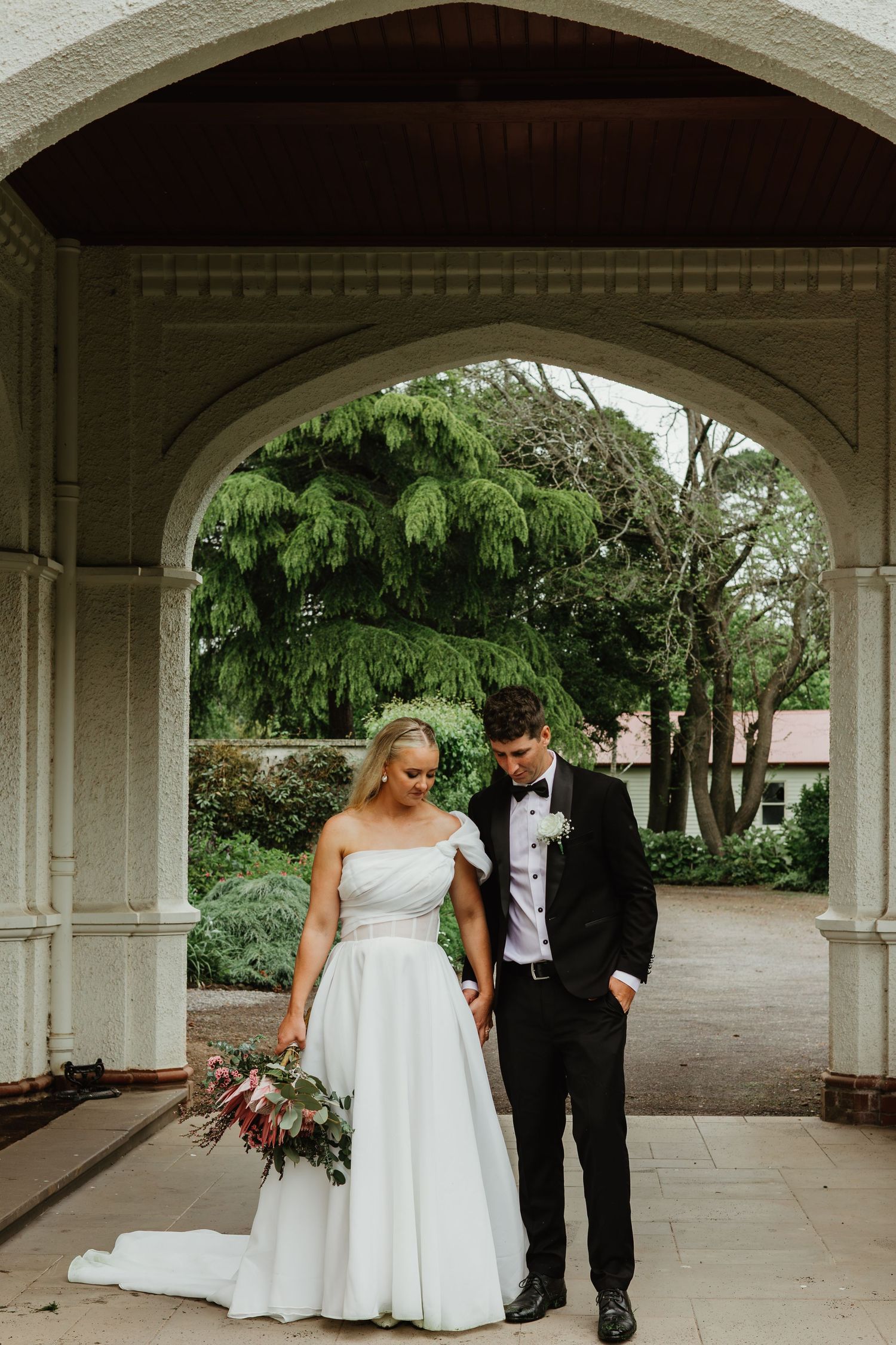 Bride and groom standing under stone arch, bride holding bouquet in white ballgown.