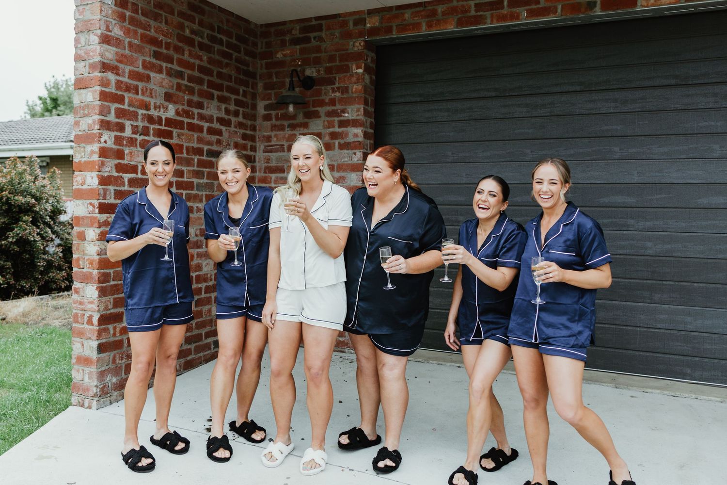 Bride and bridesmaids in matching navy satin pajamas toasting with champagne outside a brick home.