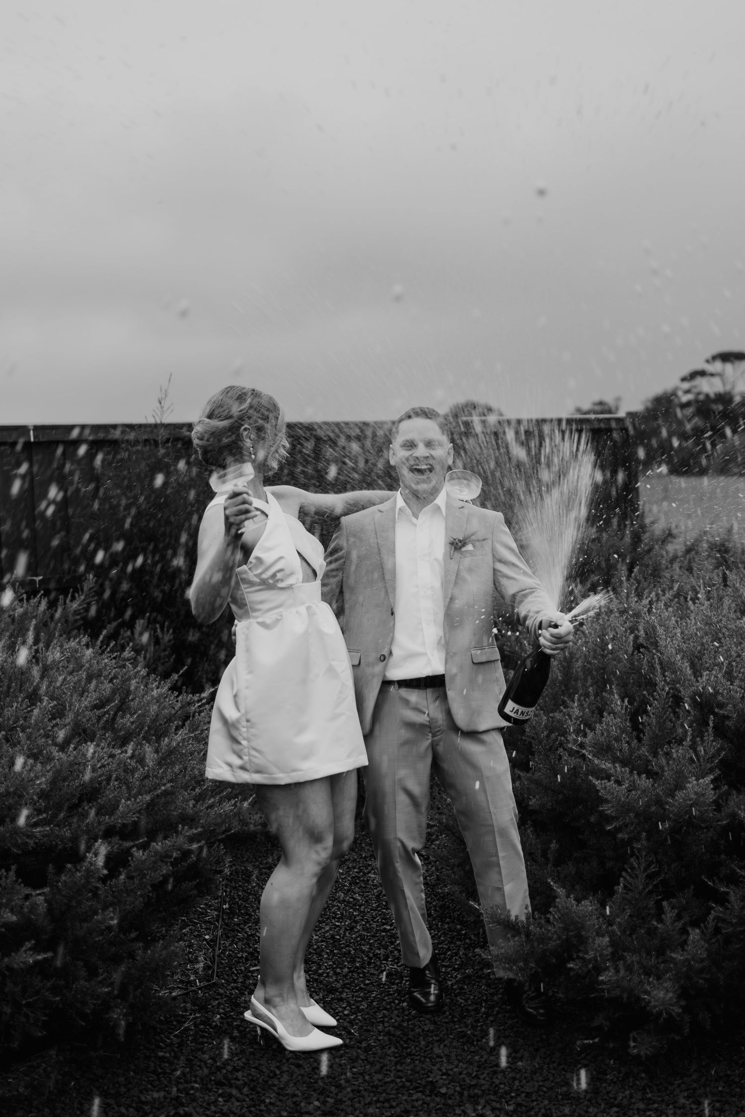 A couple joyfully pops champagne in a garden, captured in a black and white wedding photo.