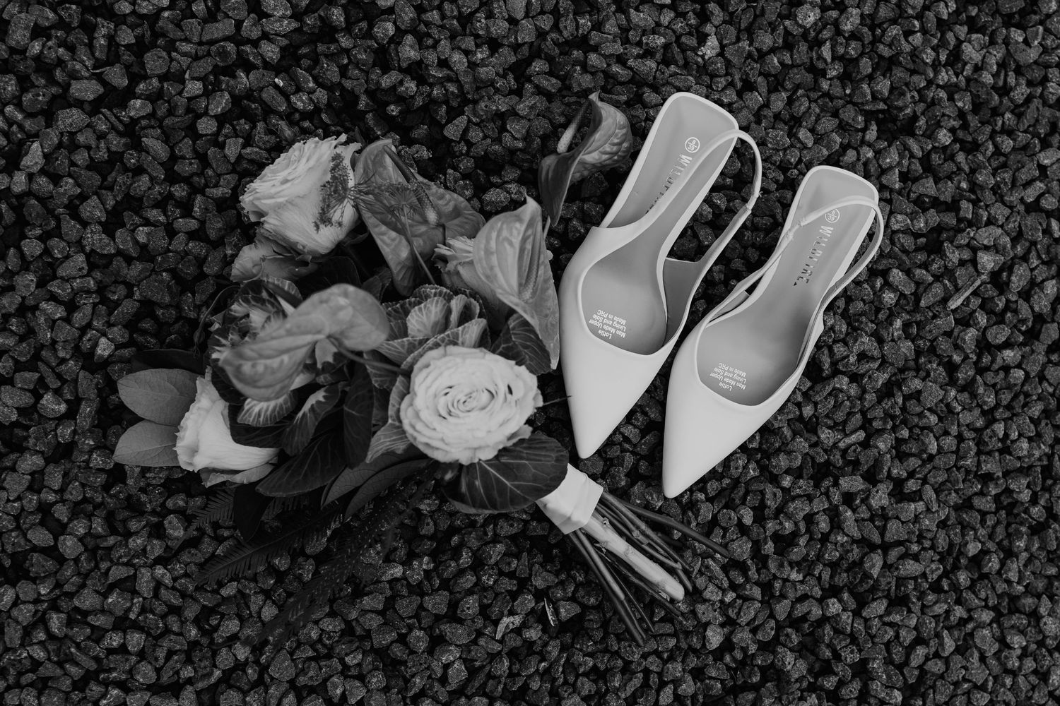 Black and white flat lay of a bridal bouquet with roses beside pointed white heels on gravel.