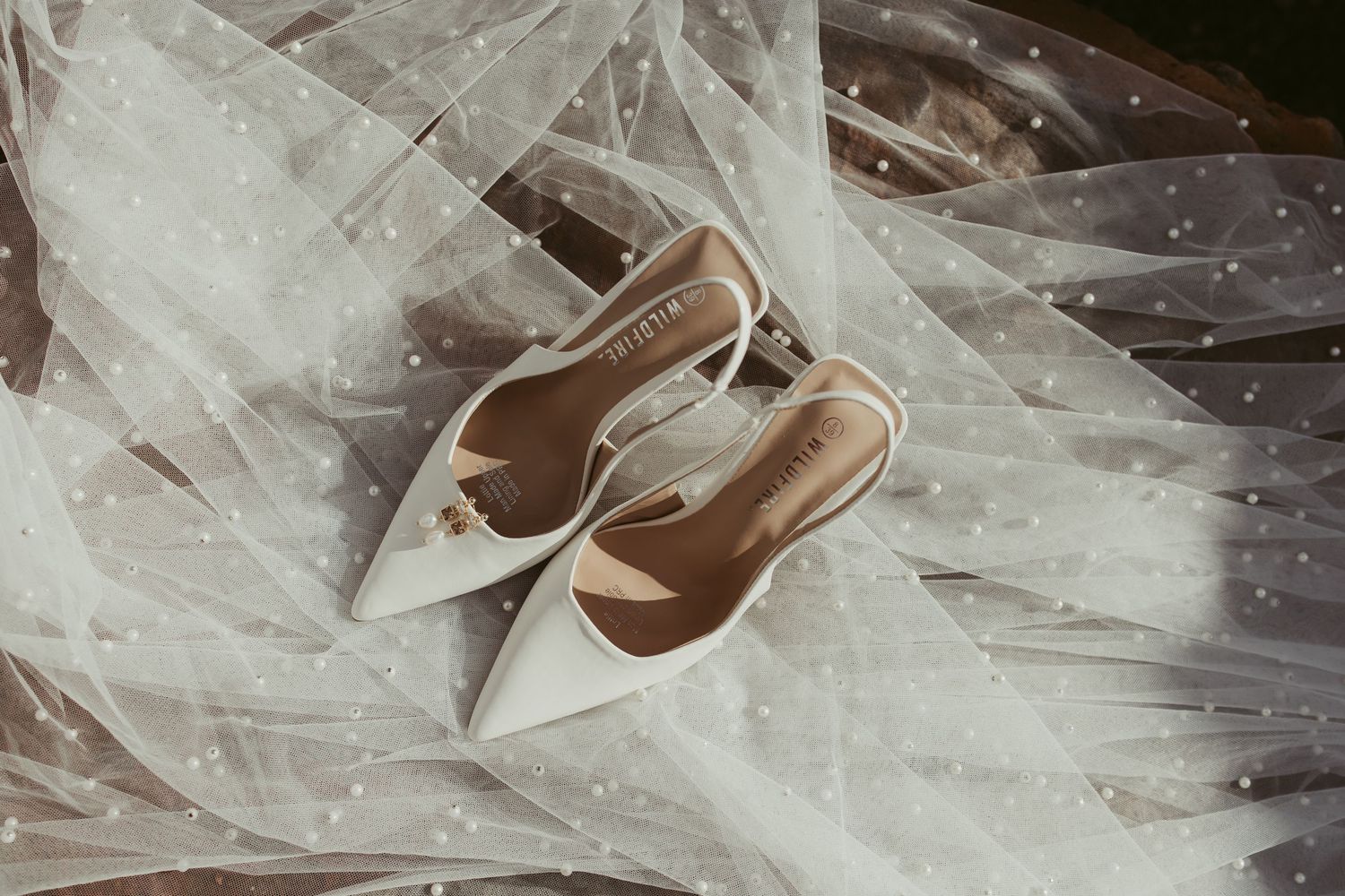 Elegant white pointed-toe slingback wedding heels placed on a delicate pearl-dotted bridal veil.
