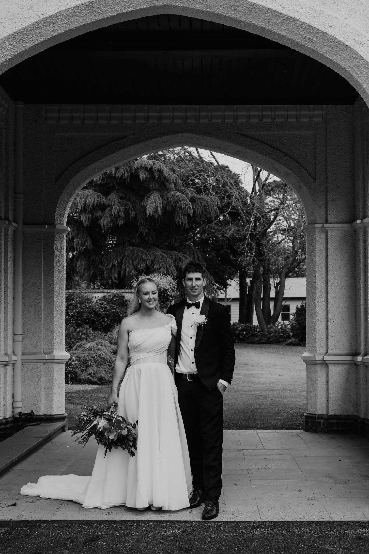 Bride and groom pose under stone archway in black and white wedding portrait surrounded by gardens.