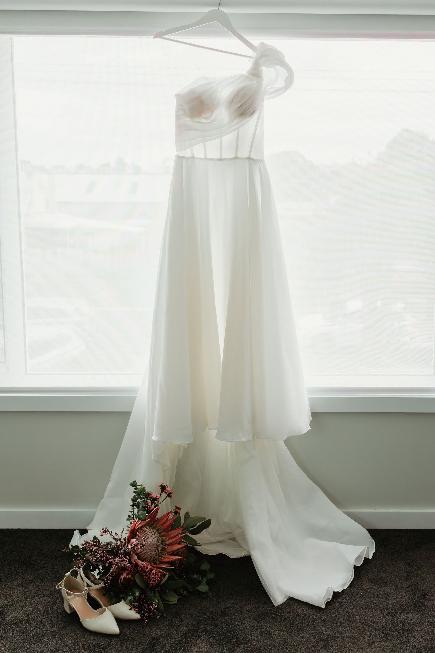 White strapless wedding gown hanging by window with bridal bouquet and heels below.