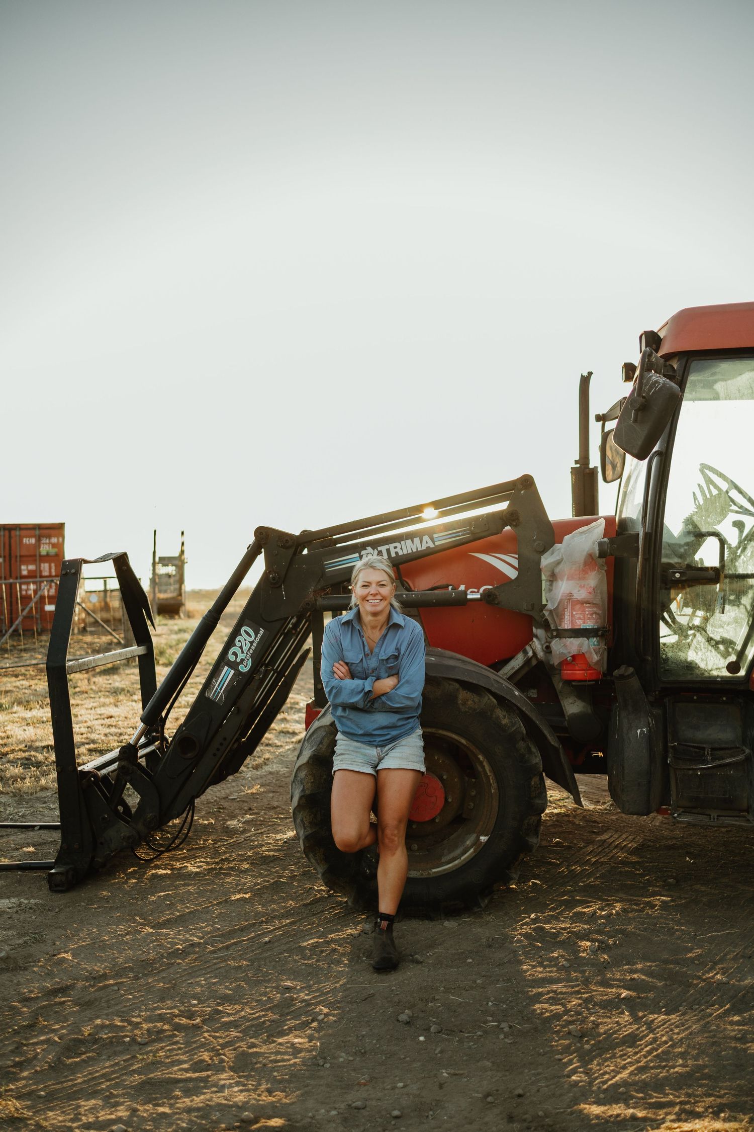Smiling woman in denim shirt leaning against red tractor on farm, arms crossed, wearing shorts and boots.