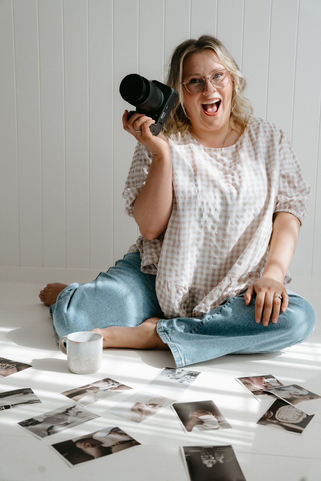 Smiling woman sitting cross-legged on white floor holding camera, surrounded by printed photos, wearing jeans and checkered blouse.