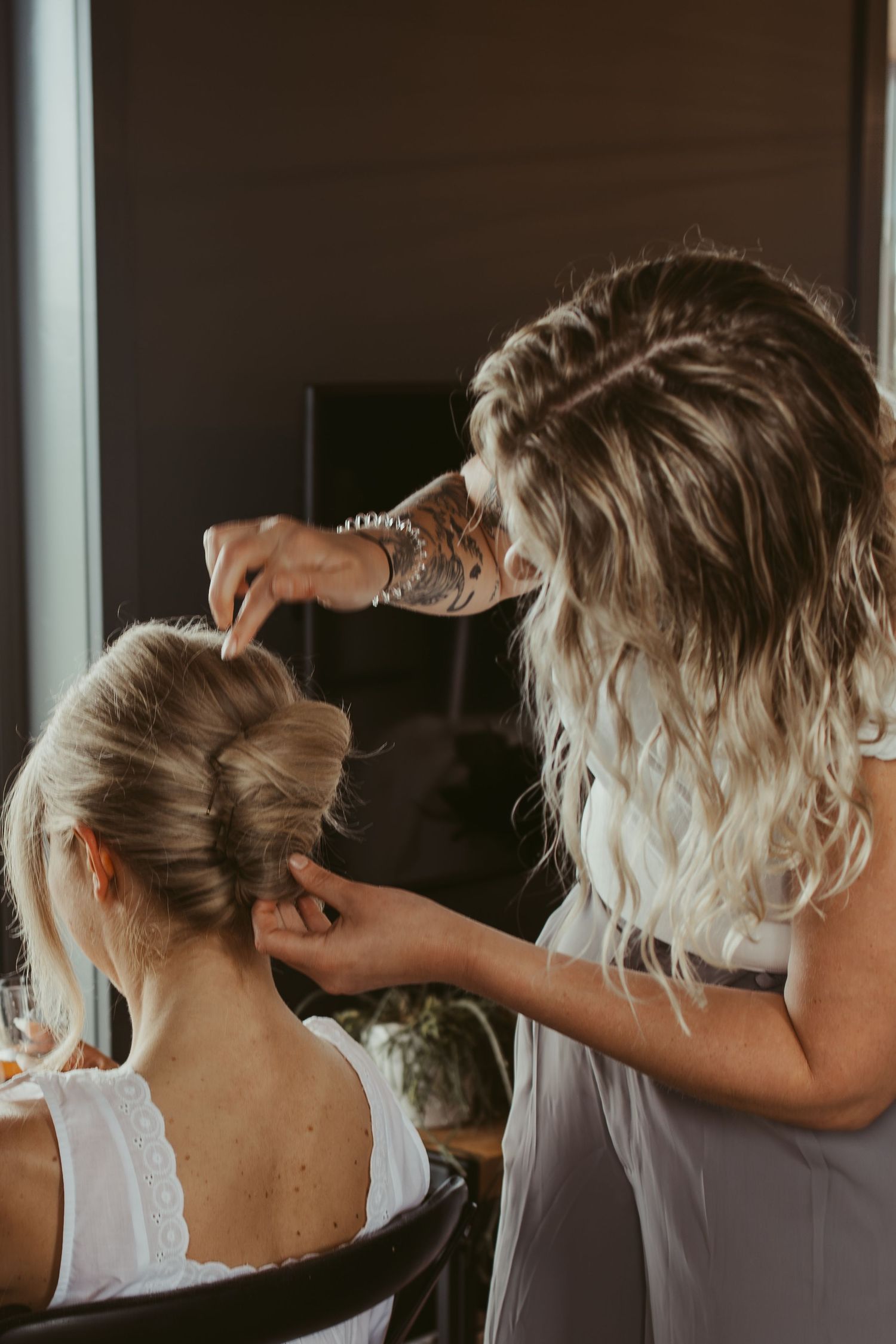 Hair stylist with tattoos styling blonde bride's elegant updo before wedding ceremony.