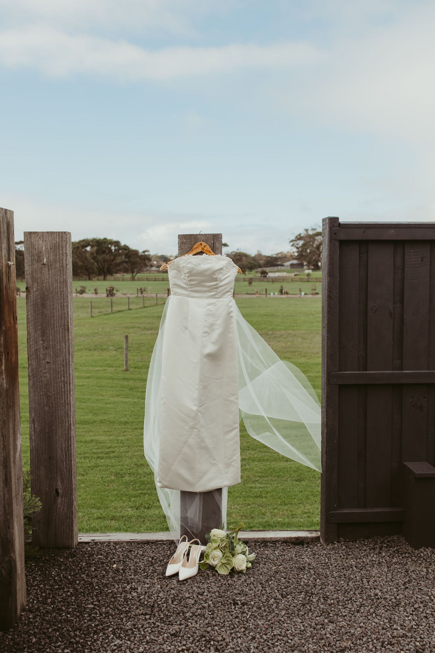 A white wedding dress with veil hanging on a wooden gate overlooking a green farm field with heels below.