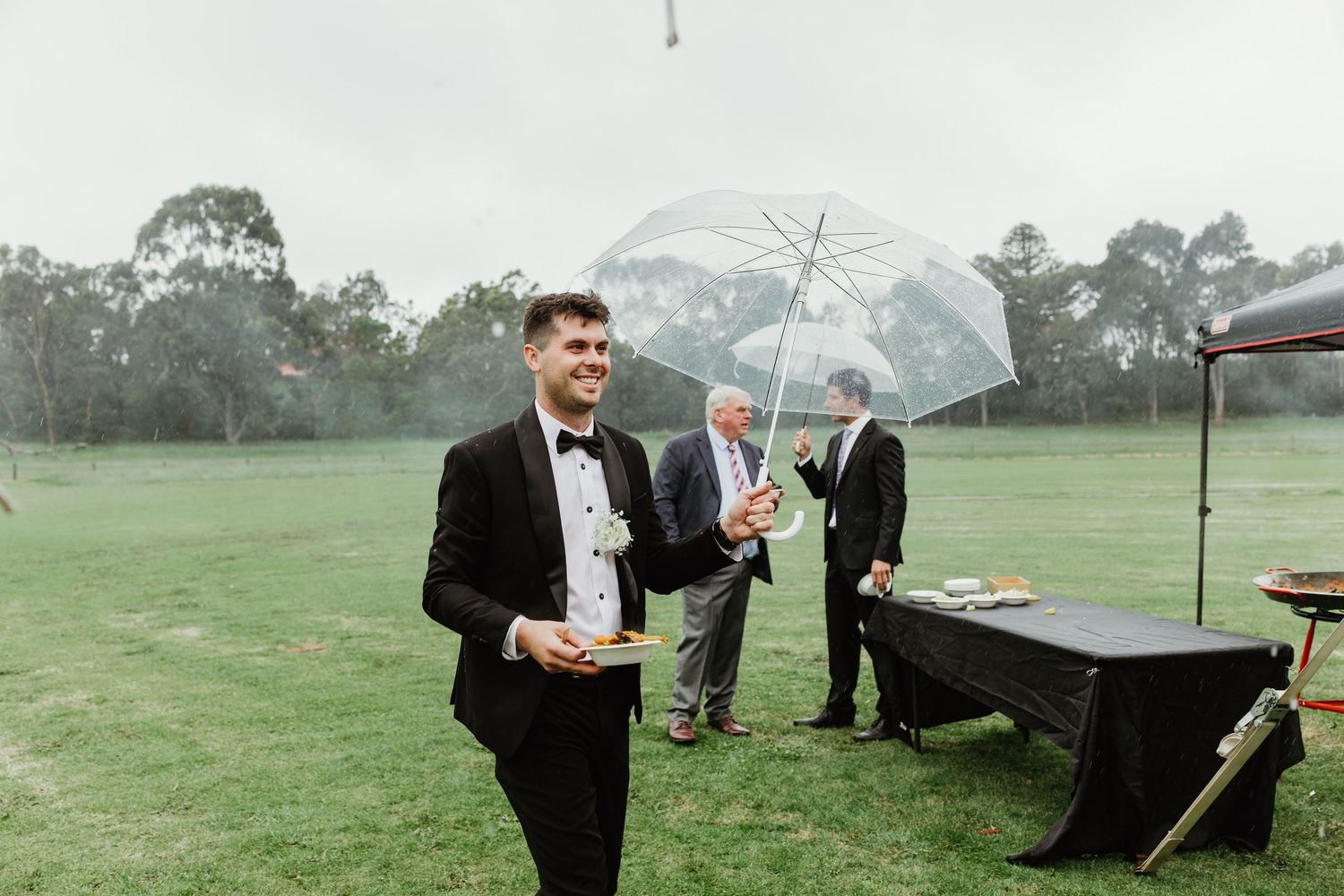 Groom in black tuxedo smiling outdoors in rain, with guests holding clear umbrella in background.