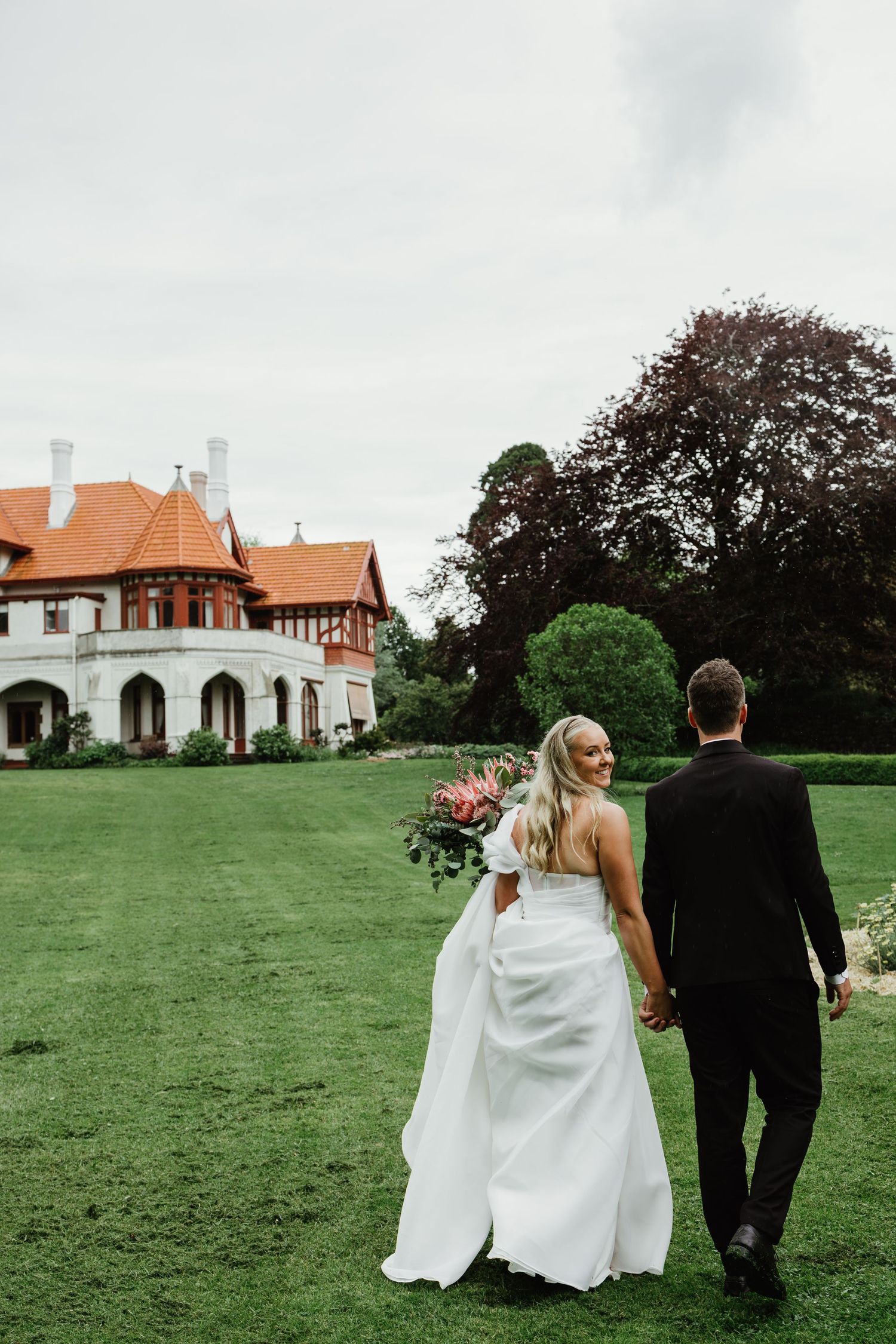 Bride and groom walking hand in hand across lush green lawn toward a grand Tudor-style wedding venue.