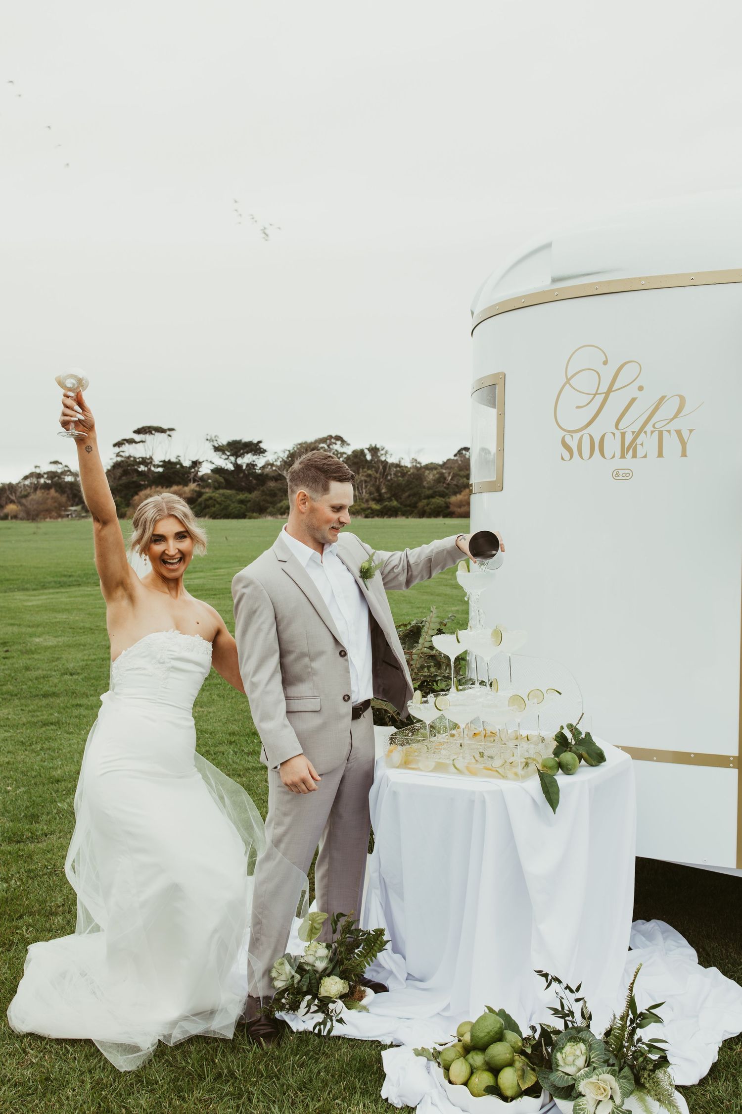 Bride and groom celebrating at Sip Society champagne tower cart on green lawn at outdoor wedding reception.