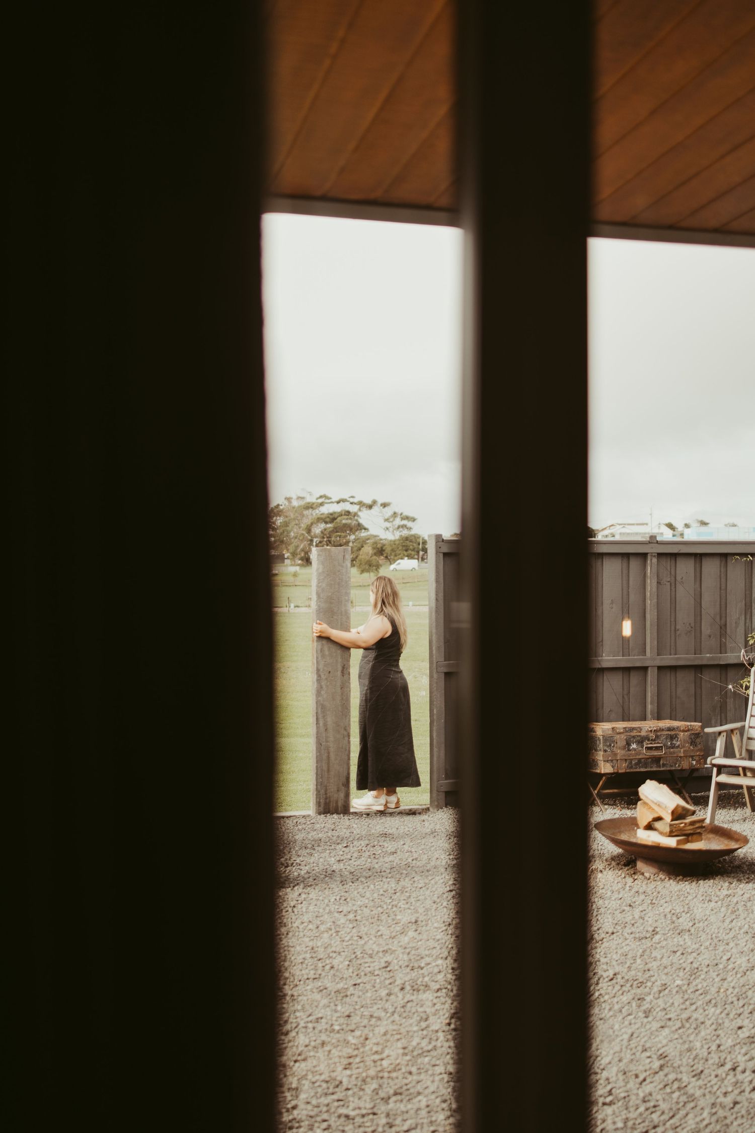 Woman in black dress viewed through window slats at rustic outdoor venue.
