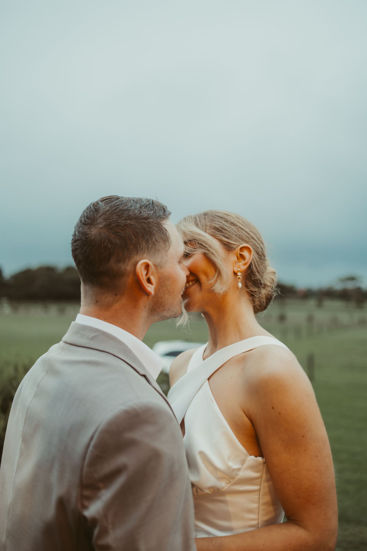 Bride and groom sharing a romantic kiss in an open field at dusk on their wedding day.