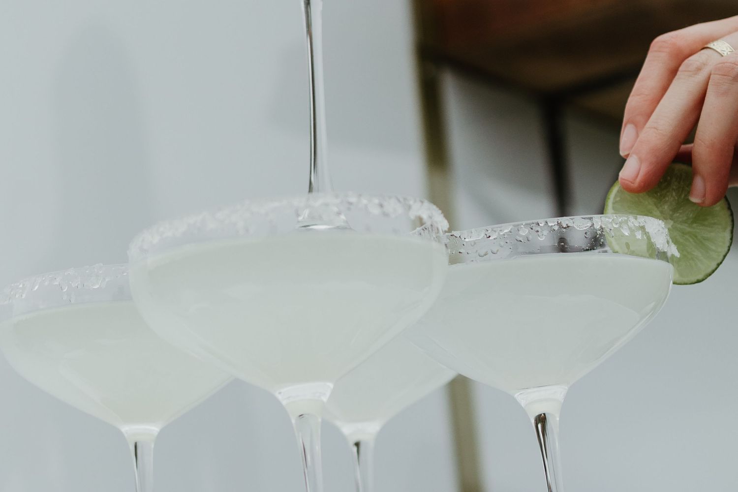 Hand garnishing margarita cocktails with lime wedge in salt-rimmed coupe glasses being poured at bar.