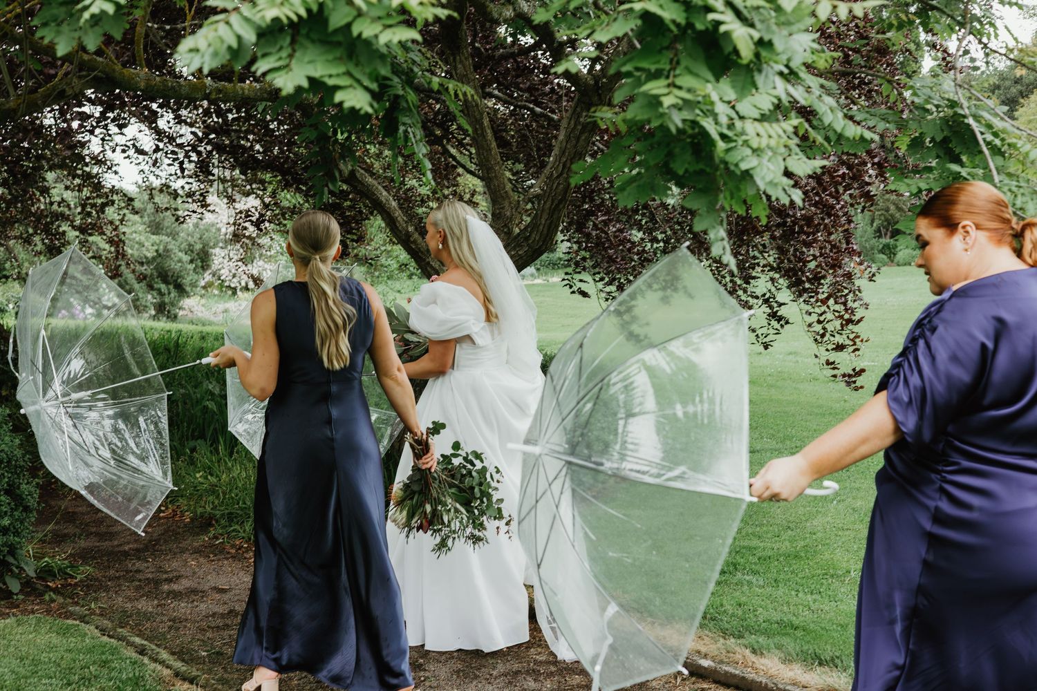 Bridesmaids in navy dresses shield bride with clear umbrellas outdoors on rainy wedding day.