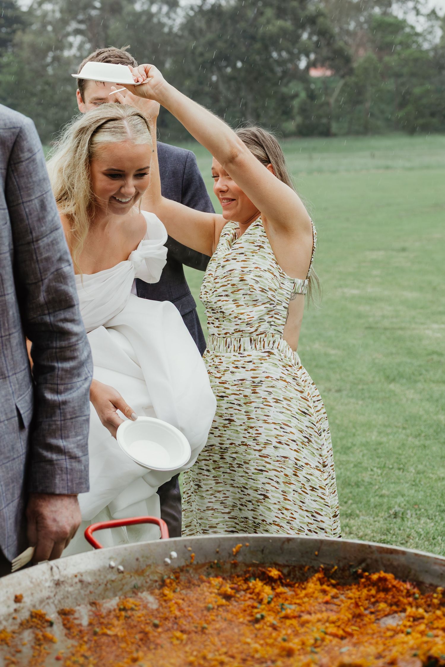Laughing bride in white gown stirs large paella pan outdoors at rustic garden wedding reception.