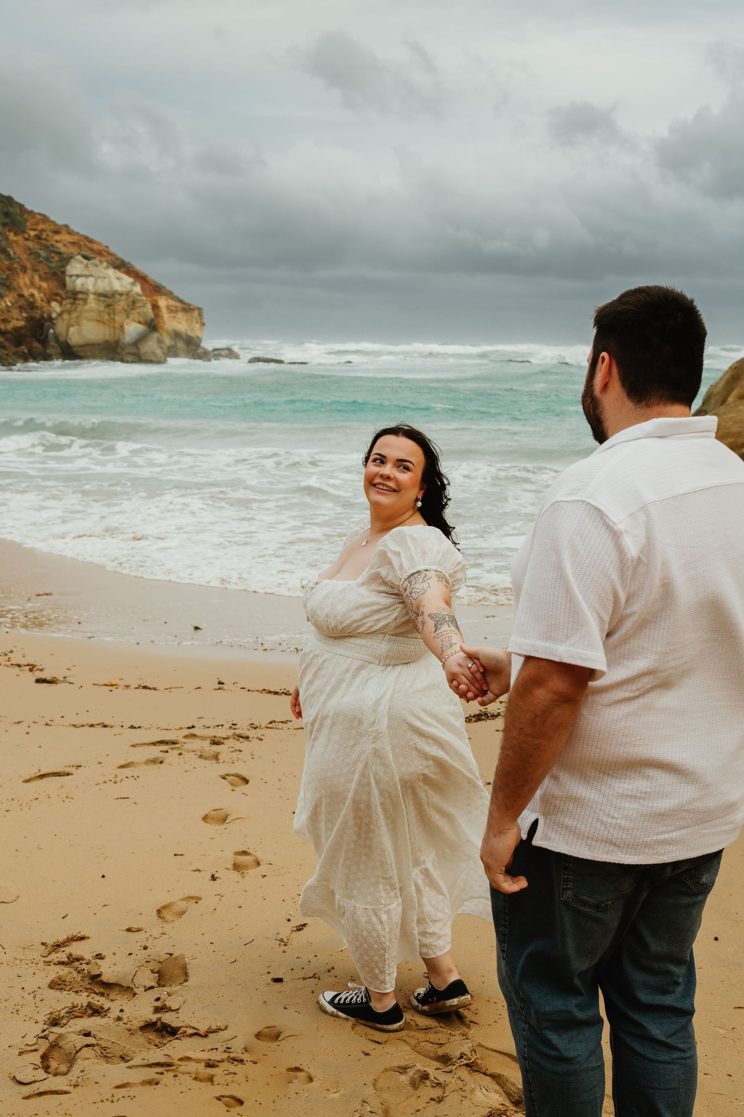 A woman in a white lace dress smiles back at her partner while walking on a stormy beach.