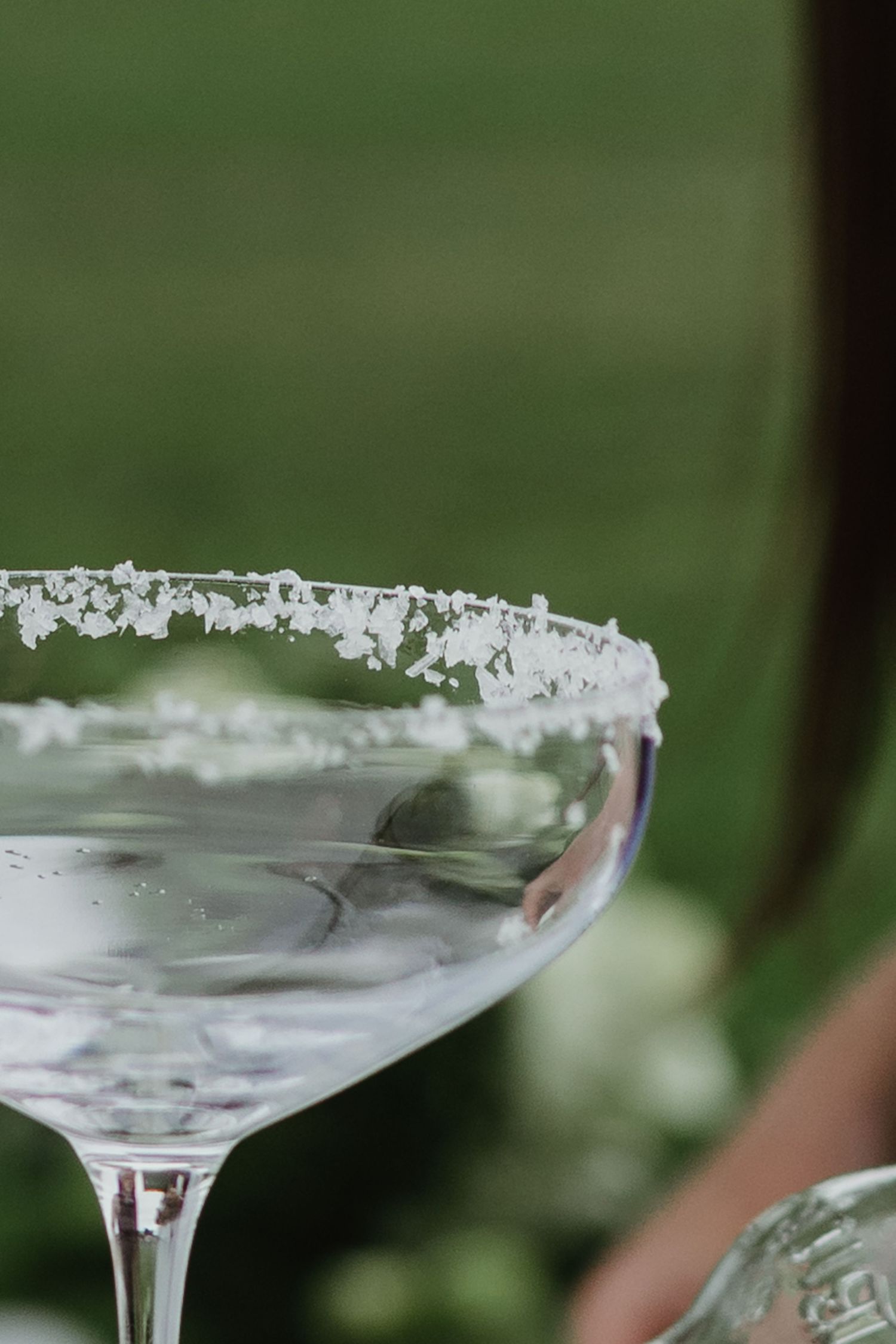 Close-up of salt-rimmed cocktail coupe glass outdoors with blurred green background.