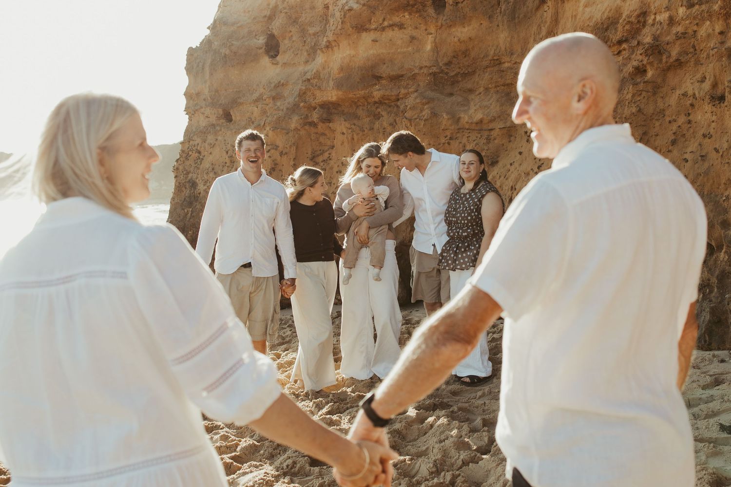 Family gathering on a rocky beach at golden hour, dressed in white and neutral tones, holding hands in a circle.