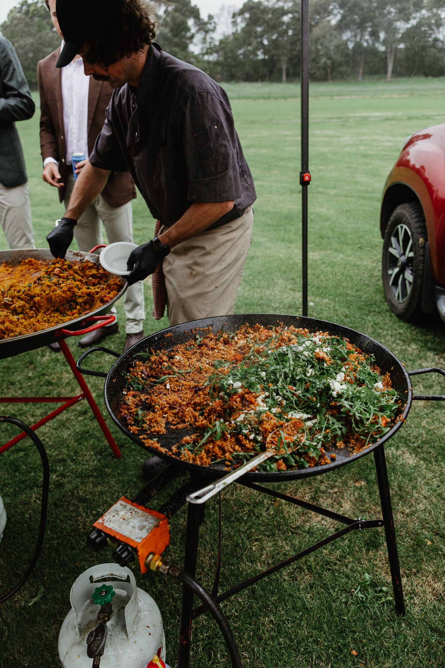 Chef serving large paella pans with vibrant rice and greens at an outdoor wedding catering station.