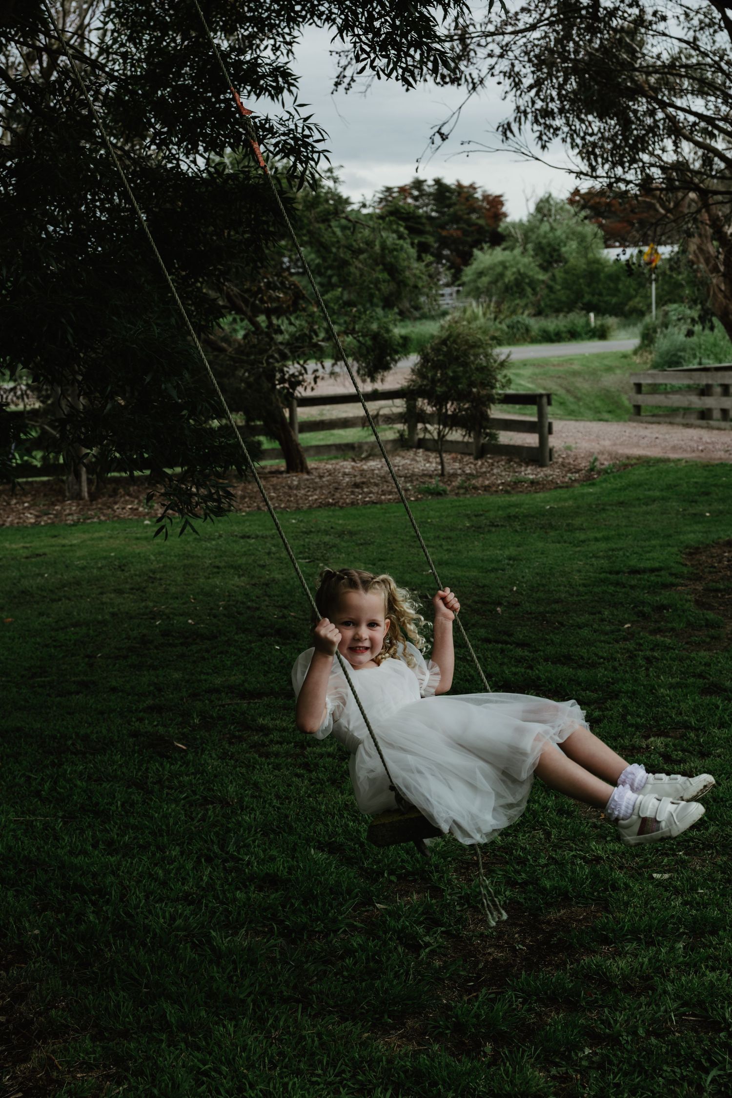 Young girl in white dress joyfully riding a rope swing in a lush green garden.