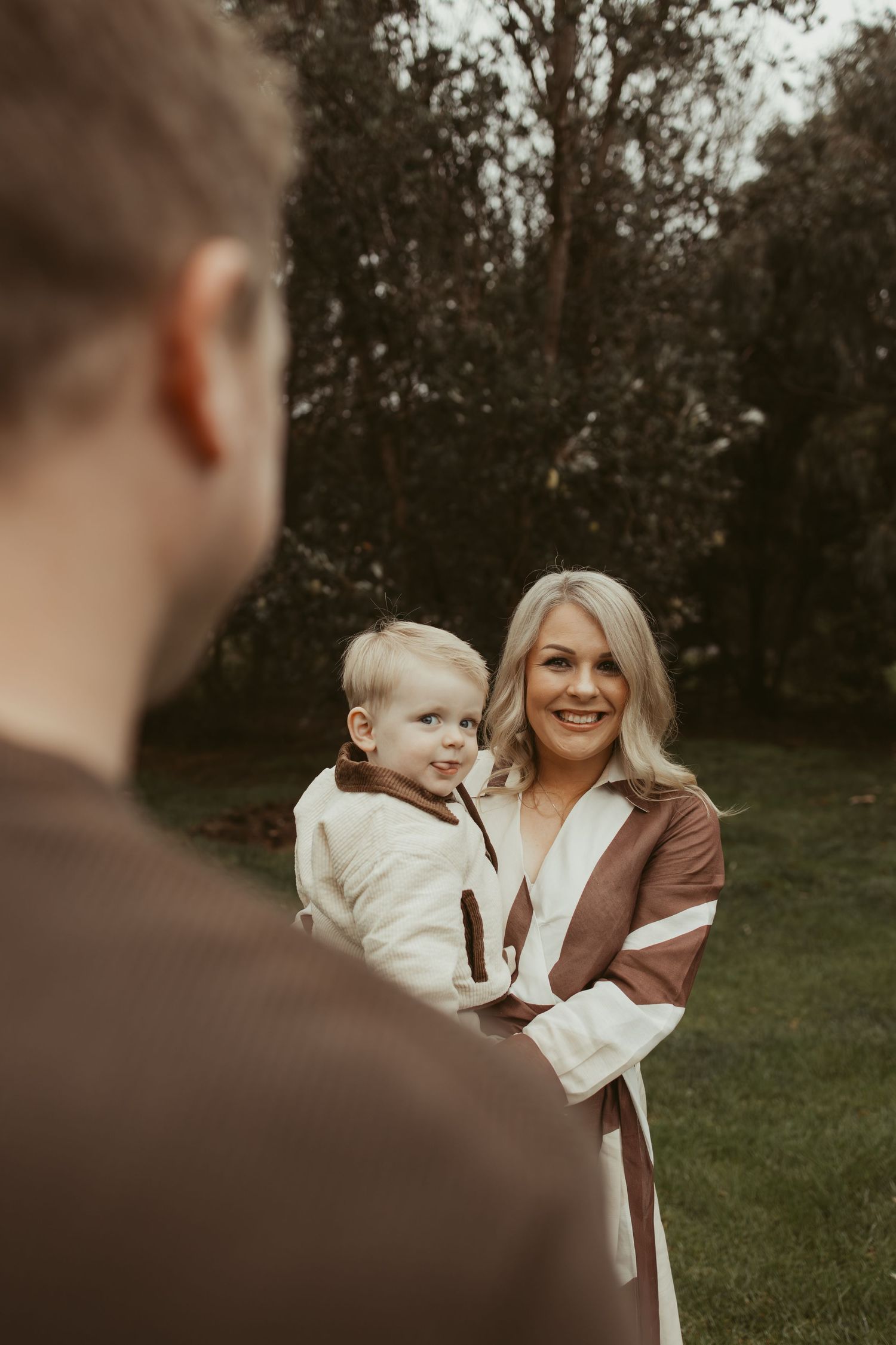 Smiling blonde mother holding toddler in white outfit while father looks on in a lush green garden.