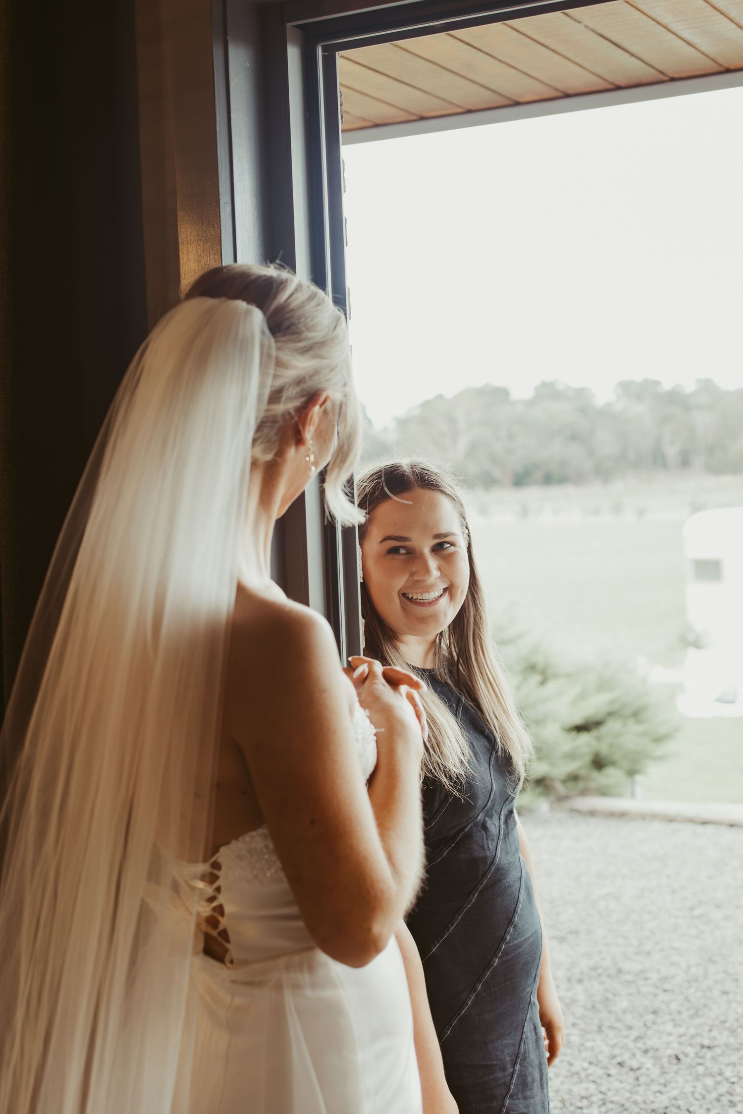 Bridesmaid smiling warmly while helping bride with wedding dress by large window.