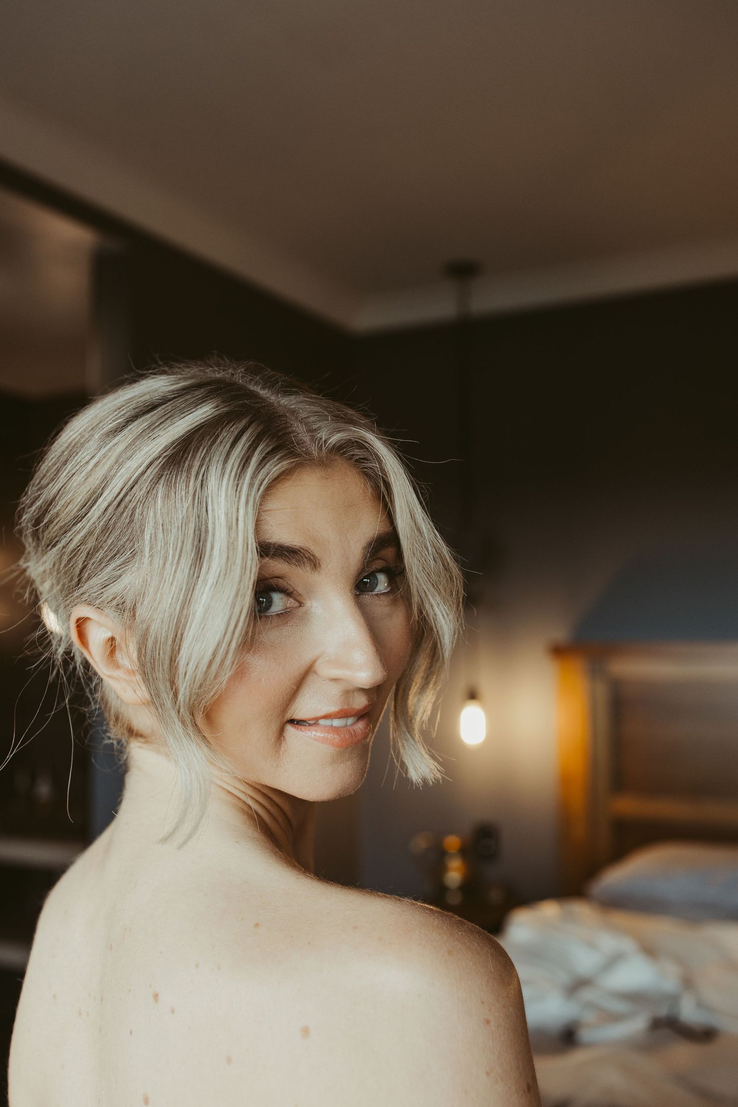 Smiling blonde bride with updo hairstyle glancing over bare shoulder in softly lit bedroom.