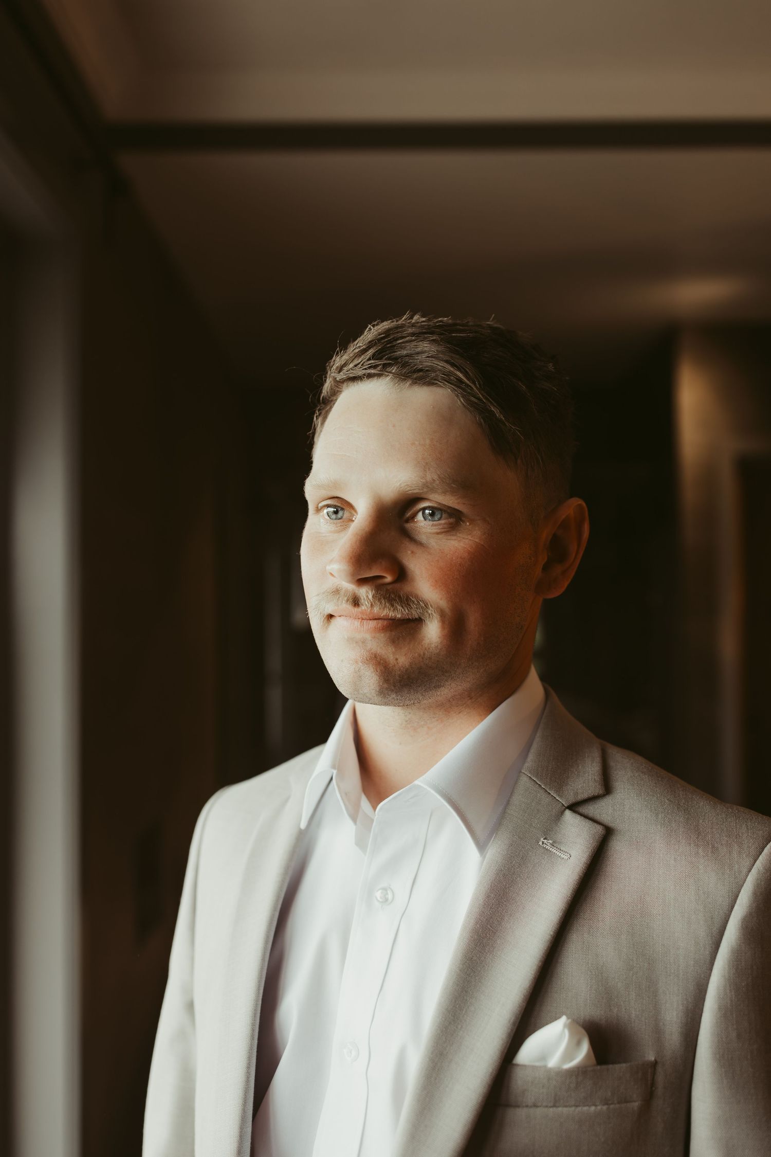 Handsome groom in light grey suit smiling softly by window on wedding day.
