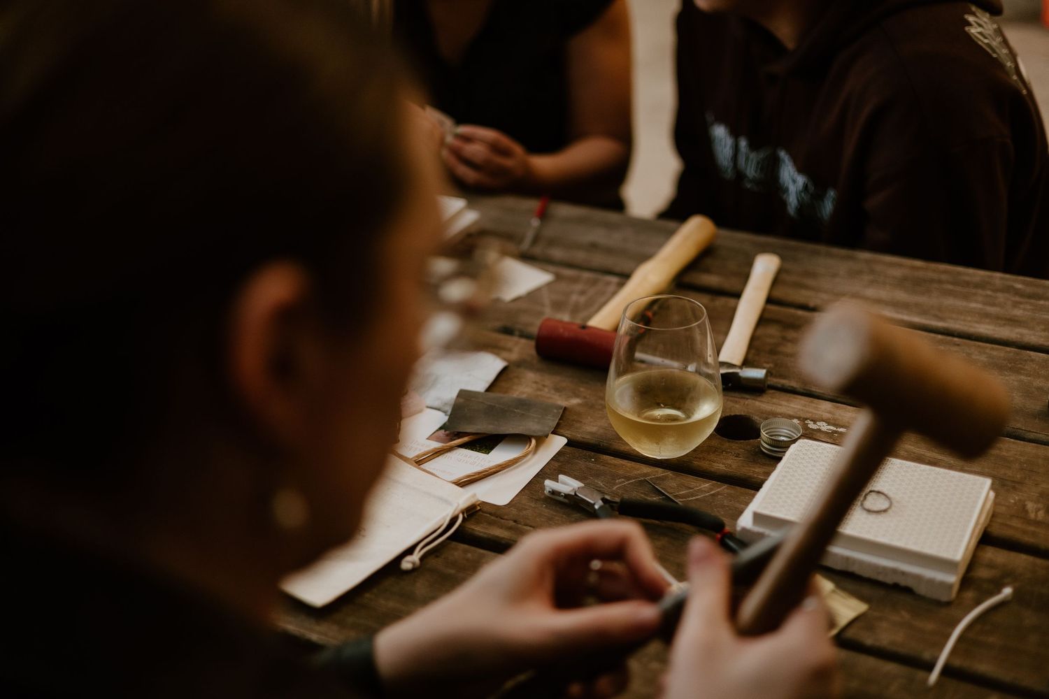 People gathered around a rustic wooden table with craft tools, leather pieces, and a glass of white wine.