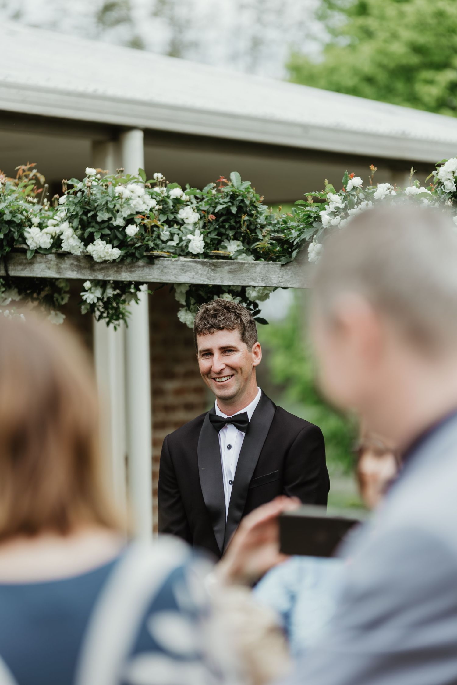 Smiling groom in black tuxedo stands at floral wedding altar with white roses and greenery.