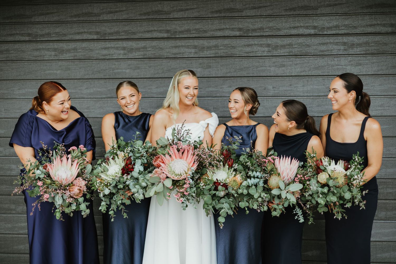 Bride and five bridesmaids in navy dresses holding large protea bouquets with greenery, smiling together against a grey wall.