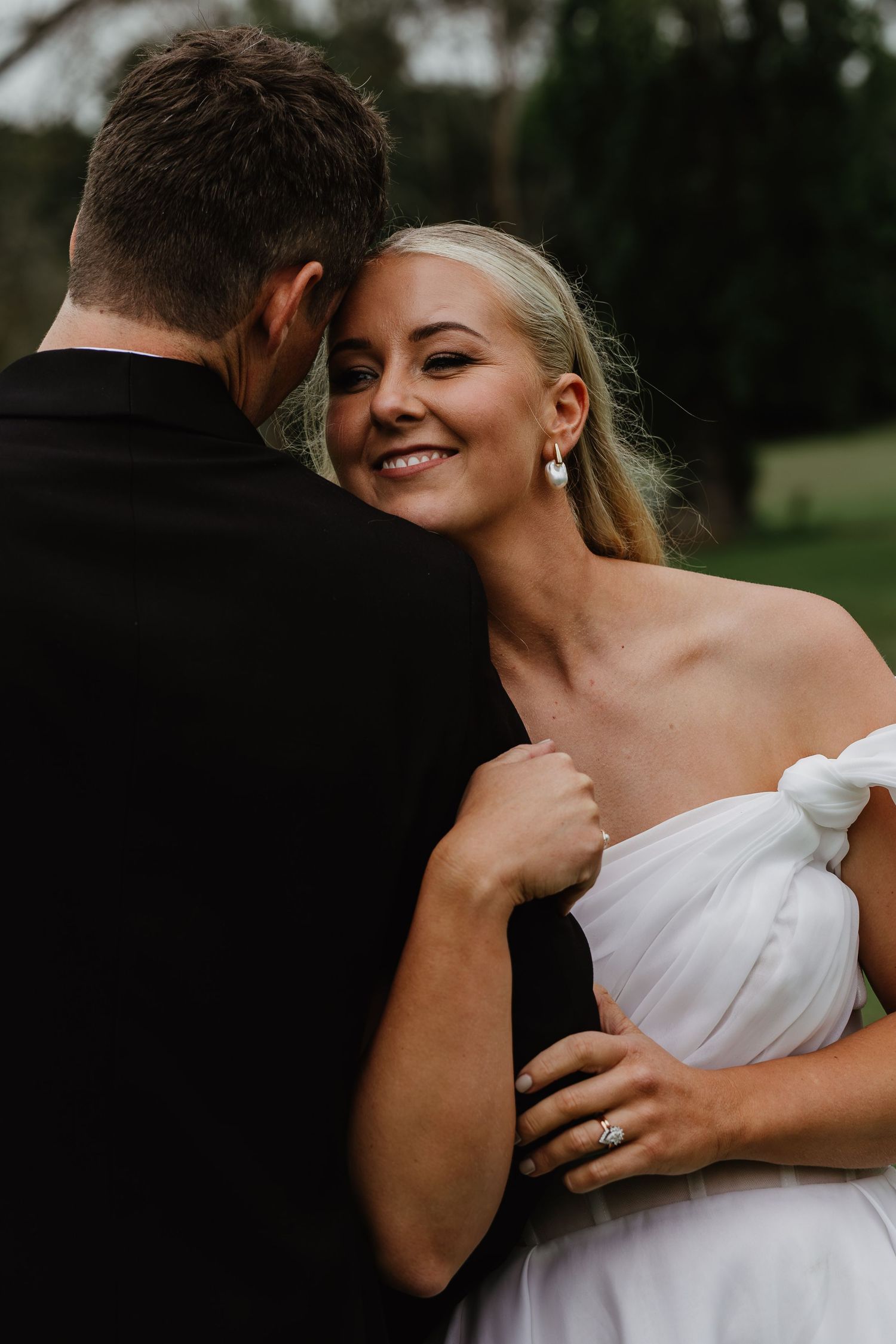 Smiling bride in white off-shoulder gown embraces groom in black tuxedo outdoors at wedding.