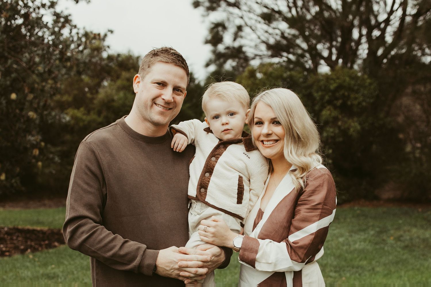 Happy family of three posing outdoors in coordinating brown and cream outfits among green trees.