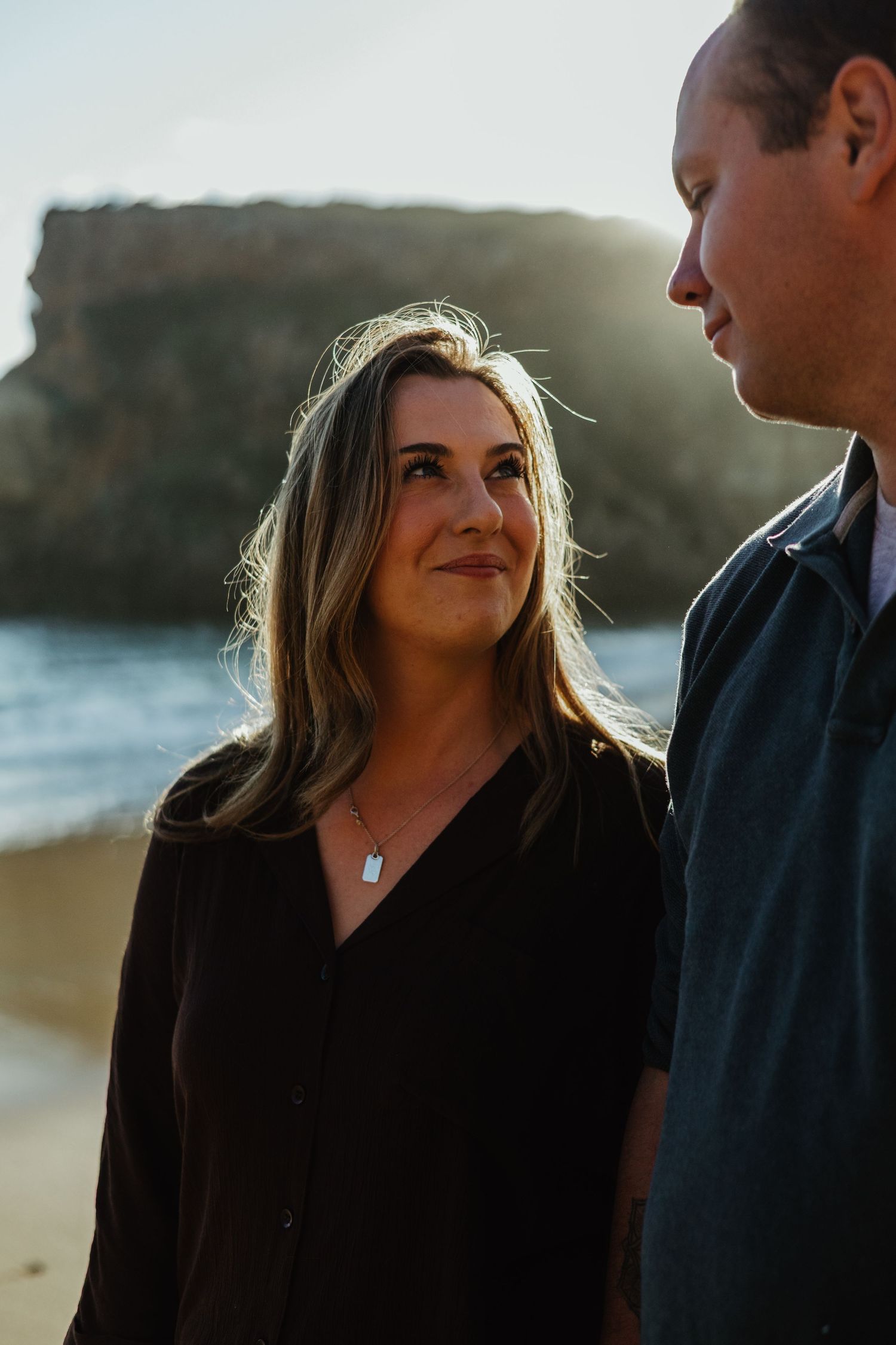 A woman with long blonde hair gazes up at a man near a rocky beach at golden hour.