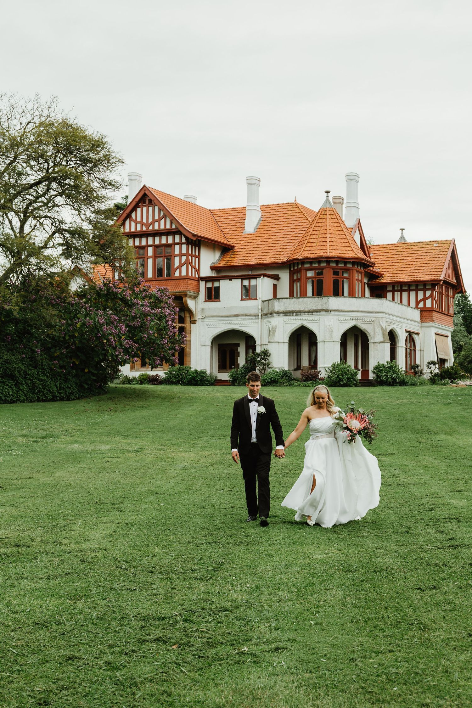 Bride and groom walk hand in hand across a green lawn before a grand Tudor-style wedding venue.