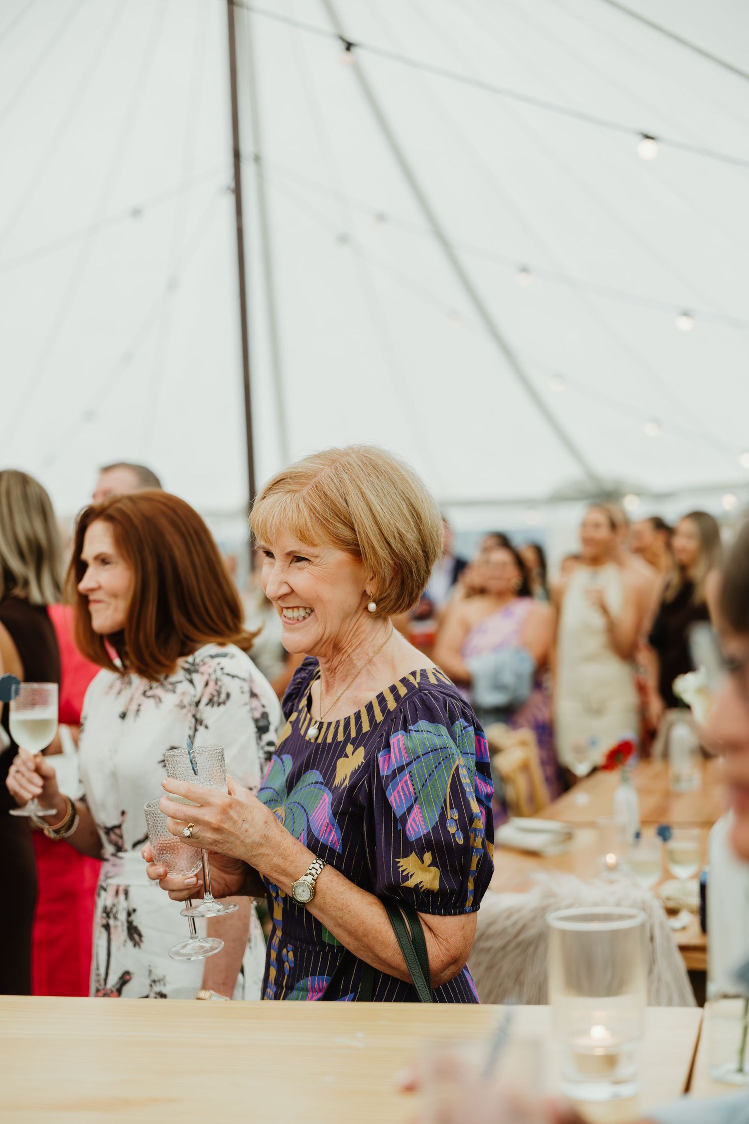 Elegant older woman in floral dress smiling and holding champagne glass at wedding reception.
