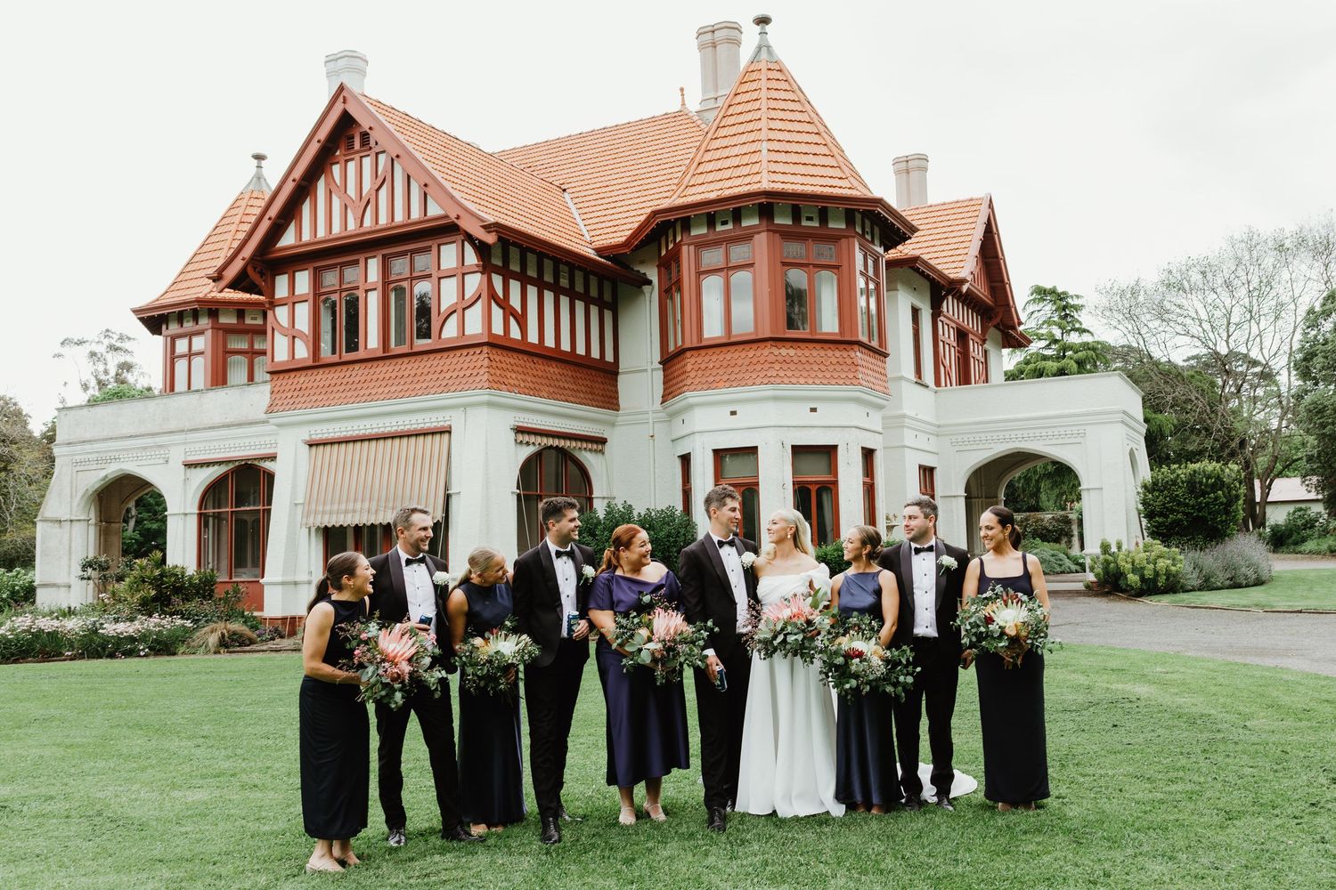 Wedding party in formal attire posing on lawn before a grand Tudor-style red and white heritage mansion.
