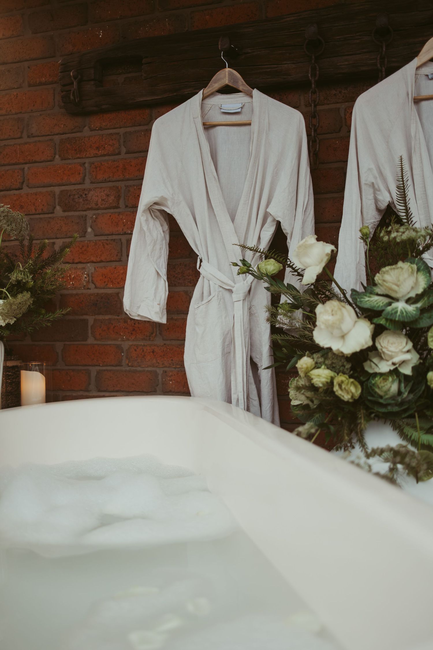 White bridal robes hanging on brick wall beside lush green and white floral arrangements.