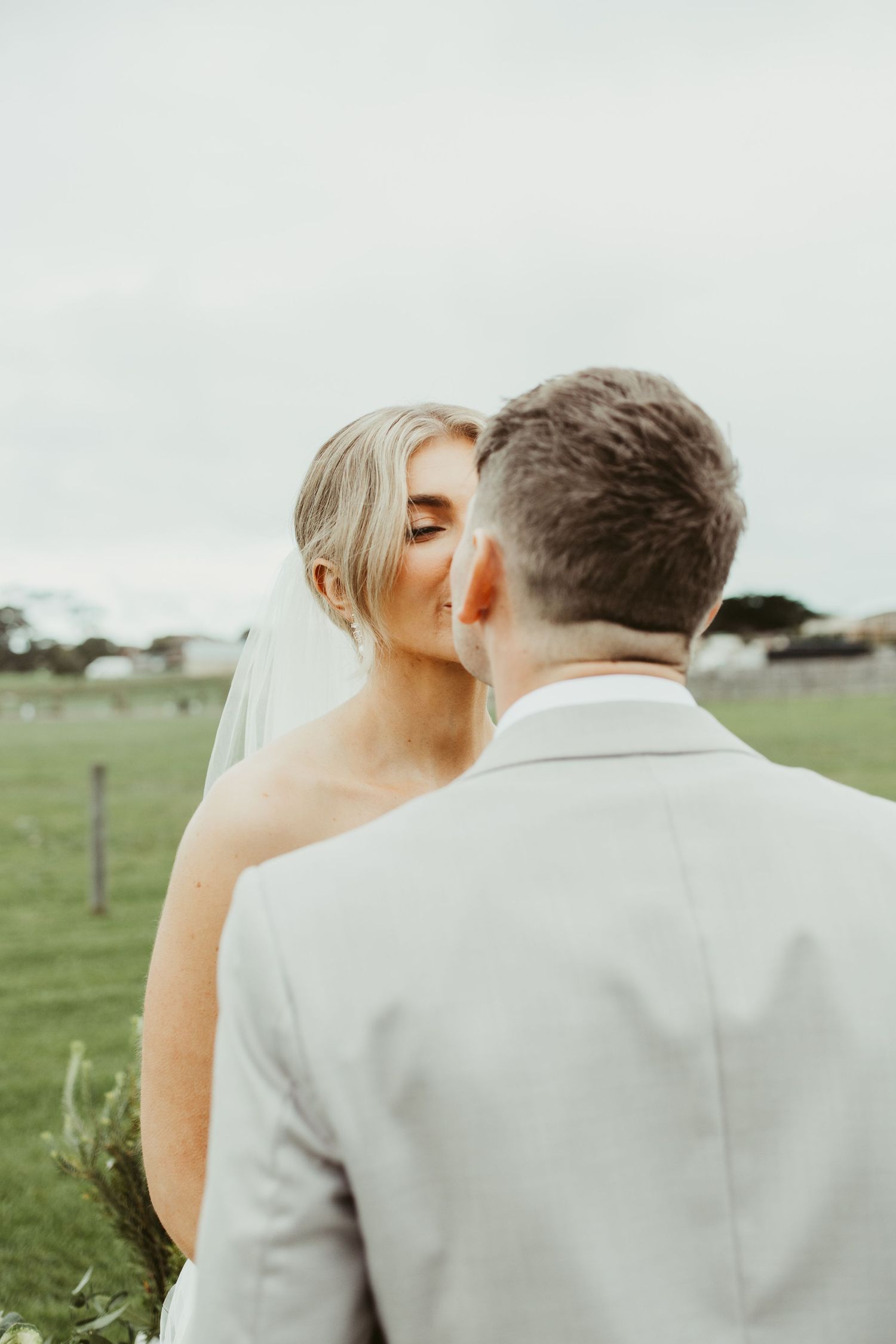 Bride kissing groom on cheek from behind in outdoor wedding setting with green field backdrop.