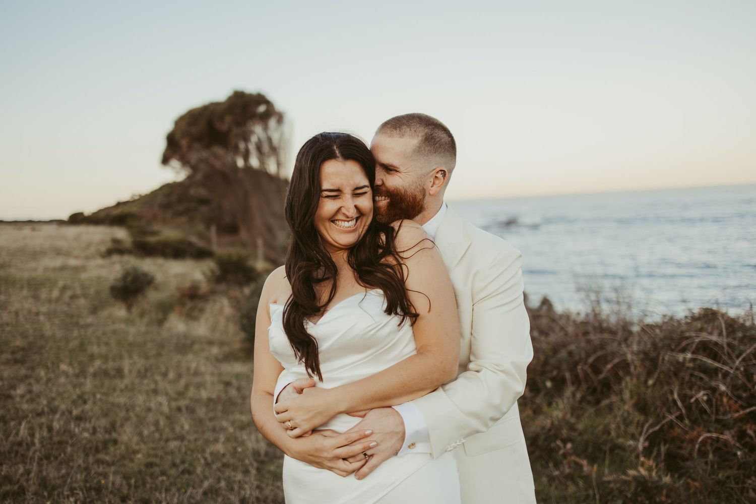 Couple embracing at golden hour on coastal cliffs, bride in white dress laughing as groom kisses her cheek.