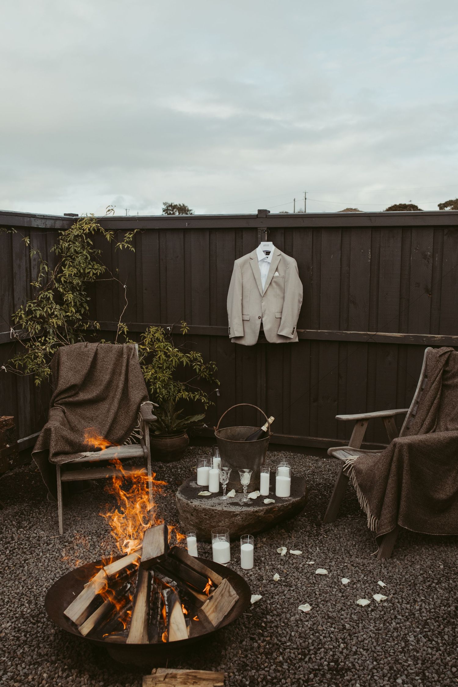 Gray suit jacket hanging on dark fence above fire pit with candles and outdoor chairs.
