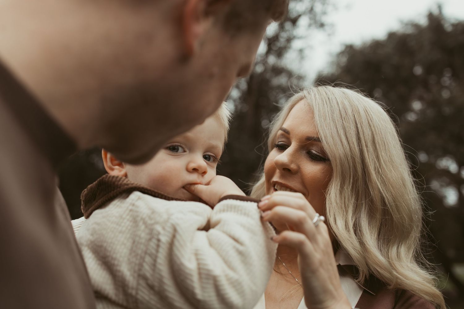 Loving mother smiling at baby held by father during outdoor family photo session.