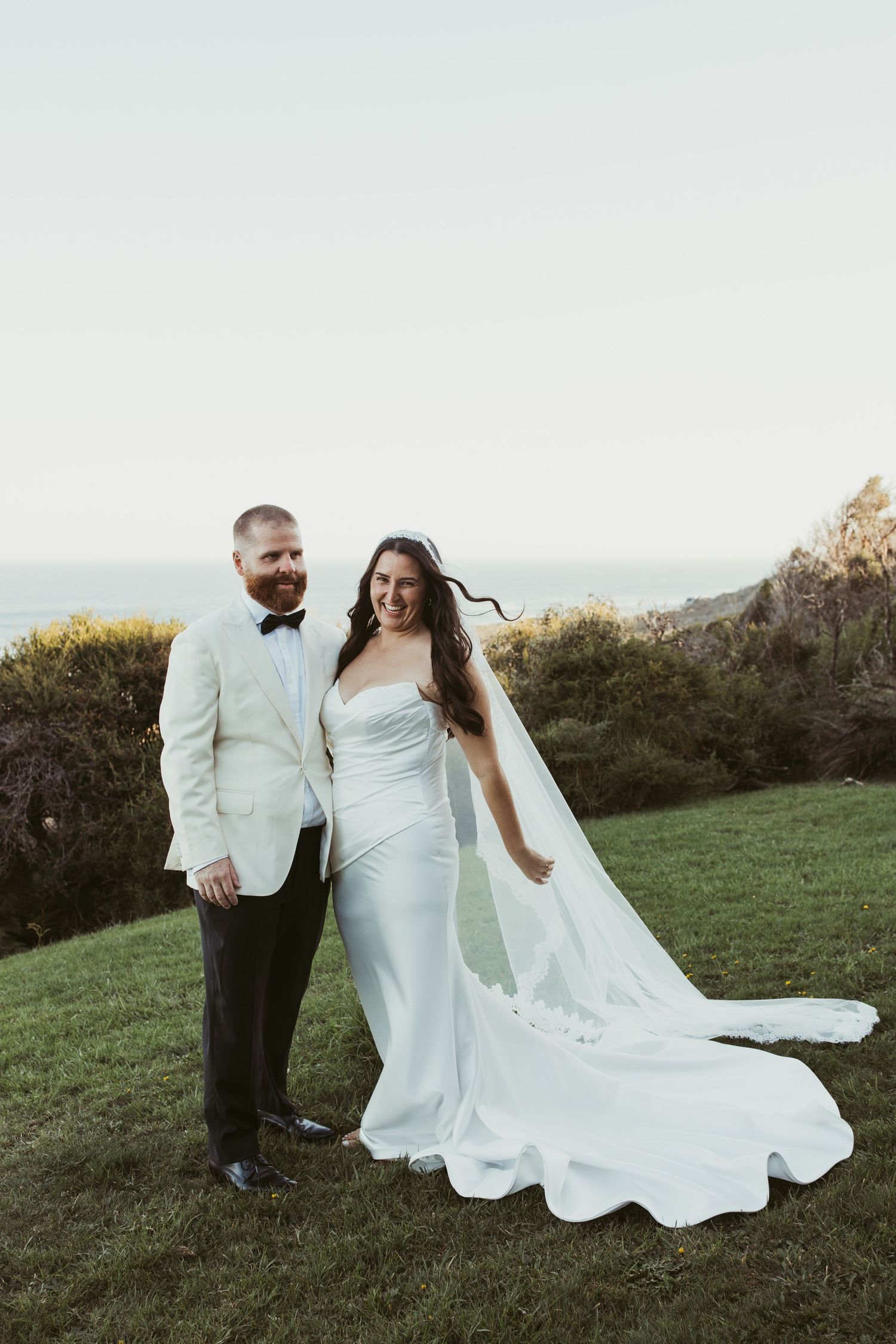 Bride and groom pose outdoors in white wedding attire with ocean cliffs backdrop on their wedding day.